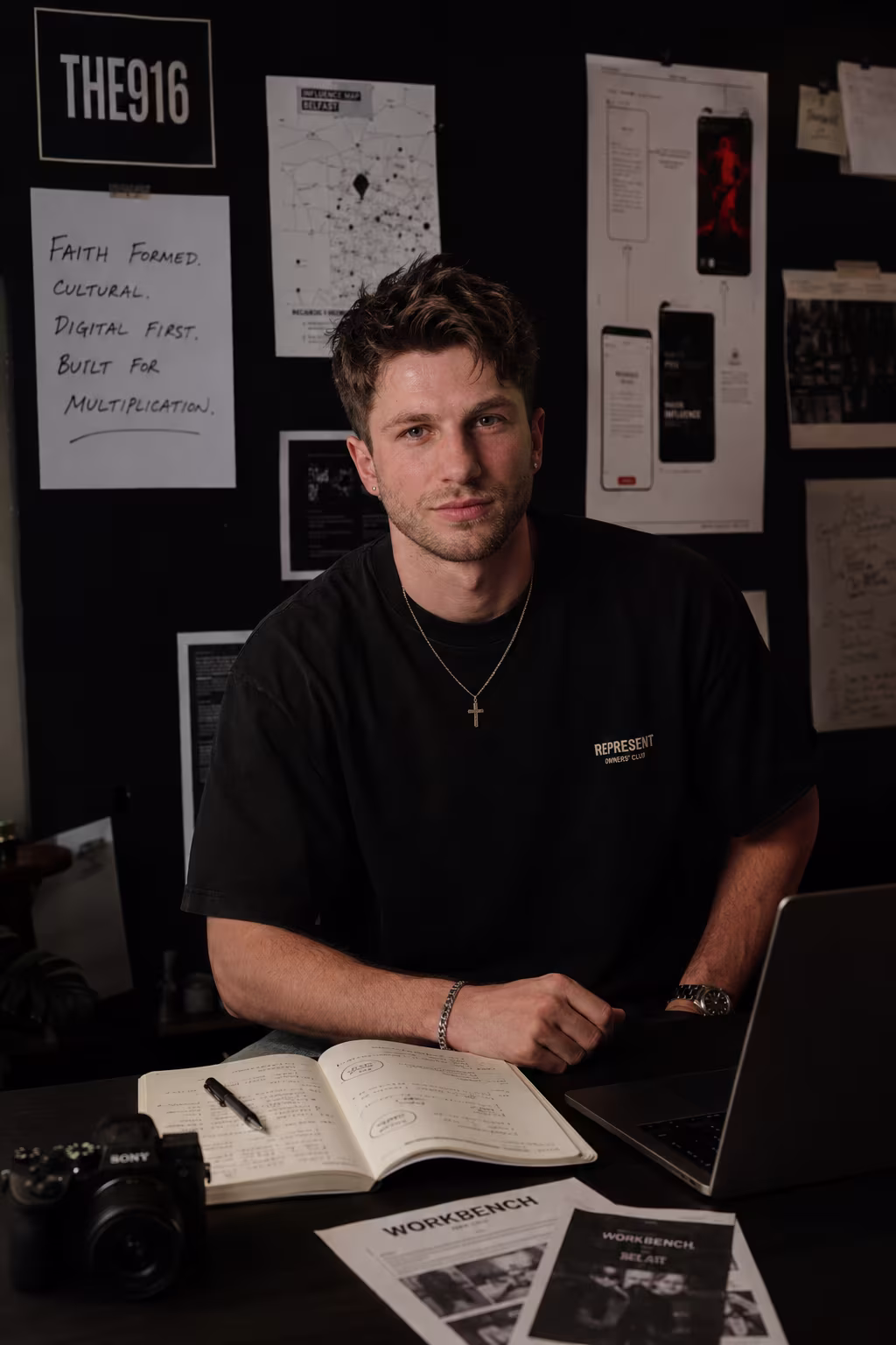 Ged Armstrong, founder of The916, at his studio desk in Belfast — a notebook, camera and laptop in front of a wall of The916 planning material and an influence map.